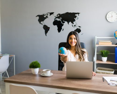 Holding the world in my hands. Attractive female travel agent showing a globe and smiling while working at her modern office